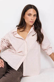 A woman wearing a light pink and white vertically striped button-down shirt, paired with brown trousers, posing against a clean white background.