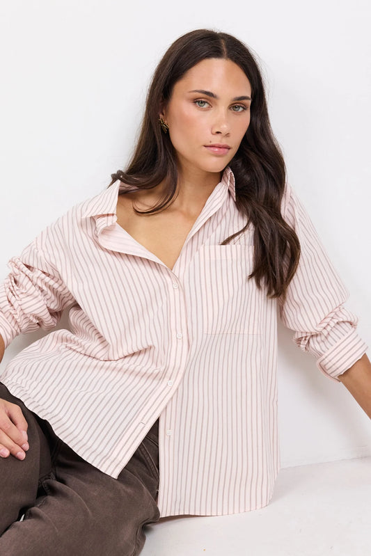 A woman wearing a light pink and white vertically striped button-down shirt, paired with brown trousers, posing against a clean white background.