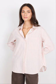 A woman modeling a light pink and white vertically striped button-down shirt with a relaxed fit, paired with dark brown pants, against a plain white studio background.