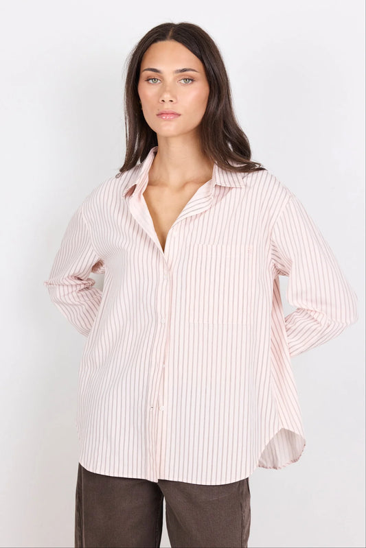 A woman modeling a light pink and white vertically striped button-down shirt with a relaxed fit, paired with dark brown pants, against a plain white studio background.