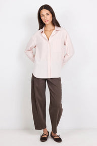 A woman modeling a light pink and white vertically striped button-down shirt with dark brown wide-leg pants and dark brown flat shoes, standing against a plain white studio background.