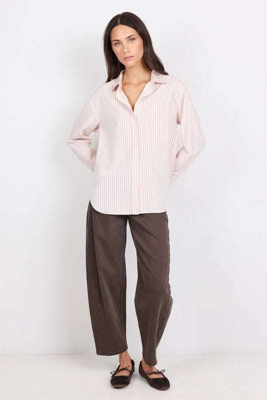A woman modeling a light pink and white vertically striped button-down shirt with dark brown wide-leg pants and dark brown flat shoes, standing against a plain white studio background.