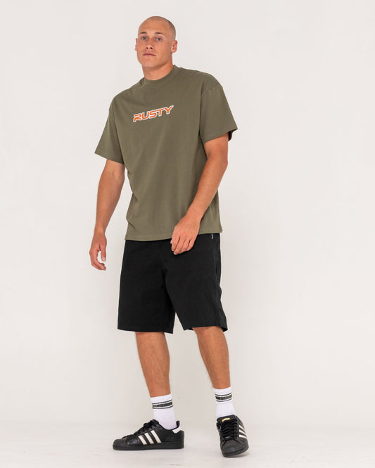 Man wearing an olive green t-shirt with 'Rusty' logo and black shorts on a white background