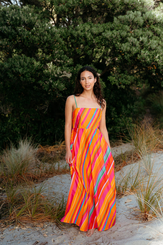 Woman wearing a colorful striped dress standing in a natural setting with greenery and sand.