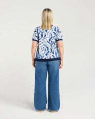 A woman viewed from the back wearing a short-sleeve top with a blue and white abstract pattern and wide-leg blue jeans, standing against a light gray studio background.