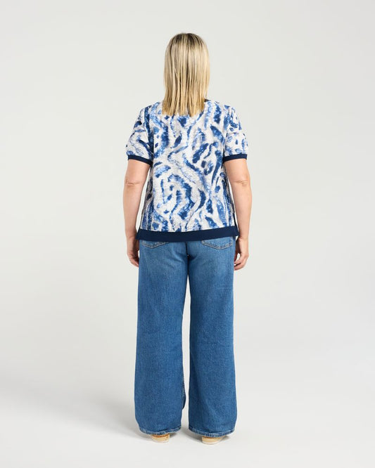 A woman viewed from the back wearing a short-sleeve top with a blue and white abstract pattern and wide-leg blue jeans, standing against a light gray studio background.