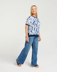 A woman modeling a short-sleeve blue and white patterned top with blue jeans and sandals, standing against a light gray studio background.