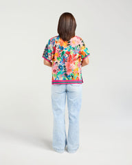 A woman viewed from behind wearing a short-sleeve tropical floral print top and light-wash flare jeans against a plain light gray studio background.