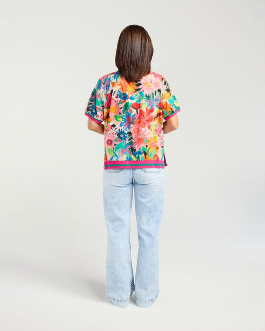A woman viewed from behind wearing a short-sleeve tropical floral print top and light-wash flare jeans against a plain light gray studio background.