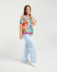 A woman modeling a colorful short-sleeve t-shirt with a vibrant floral pattern, paired with light-wash wide-leg jeans and white sneakers, against a plain light gray studio background.