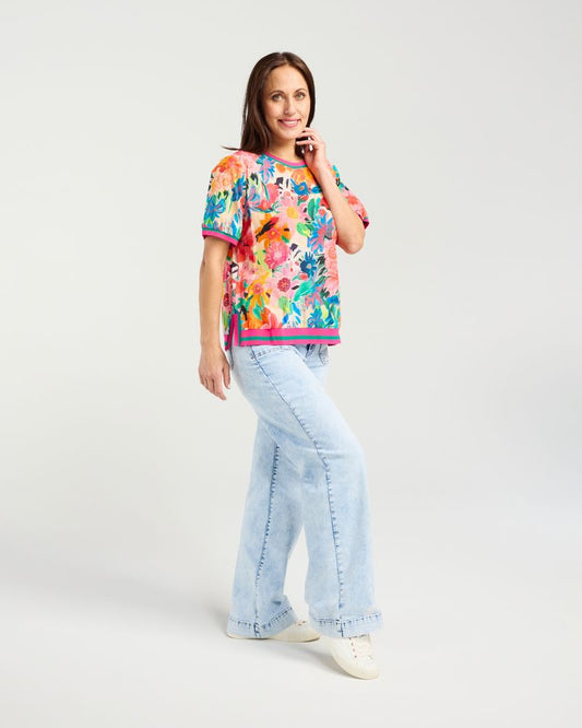 A woman modeling a colorful short-sleeve t-shirt with a vibrant floral pattern, paired with light-wash wide-leg jeans and white sneakers, against a plain light gray studio background.