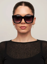 Woman wearing a black outfit and sunglasses against a plain background