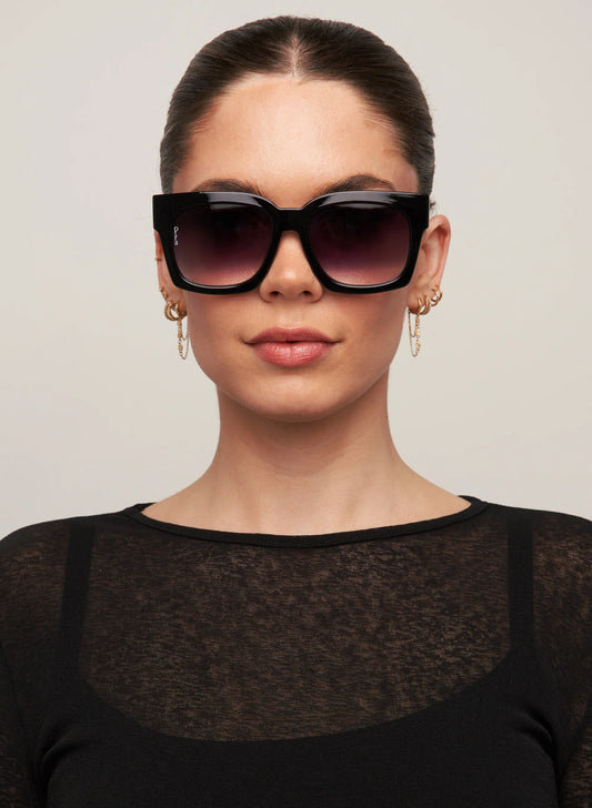 Woman wearing a black outfit and sunglasses against a plain background