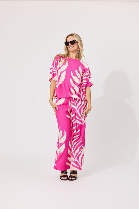 A woman modeling a bright pink and white palm leaf print two-piece set consisting of a short-sleeve top and matching wide-leg pants, styled with black sunglasses and black sandals against a plain white studio background.