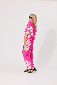A woman modeling a bright pink and white abstract patterned jumpsuit, styled with black sunglasses and black heeled sandals, standing against a plain white studio background.
