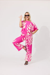 A woman modeling a bright pink and white tropical leaf print two-piece set consisting of a short-sleeve top and matching wide-leg pants, styled with black sunglasses and black sandals against a plain white studio background.