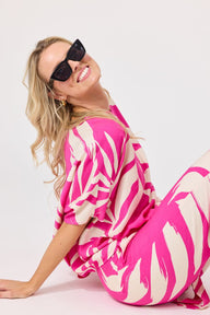A woman wearing a bright pink and cream abstract print kaftan dress and matching trousers, styled with black sunglasses, posing on a light gray studio background.