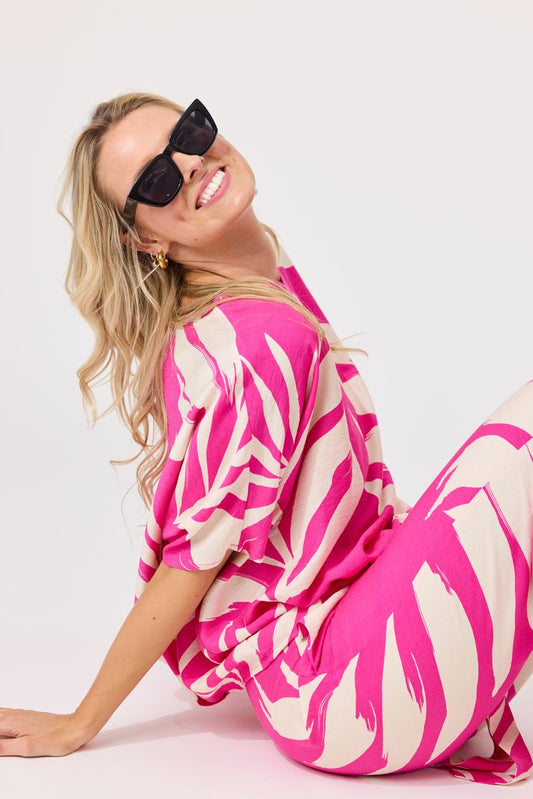 A woman wearing a bright pink and cream abstract print kaftan dress and matching trousers, styled with black sunglasses, posing on a light gray studio background.