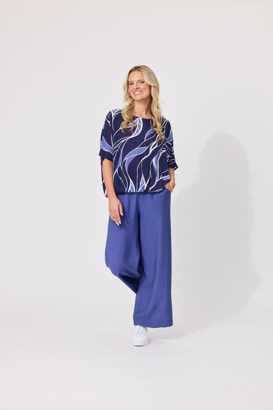 A woman modeling a two-piece outfit consisting of a navy blue short-sleeve top with a white and light blue abstract wave pattern, paired with wide-leg royal blue pants, against a plain light gray studio background.