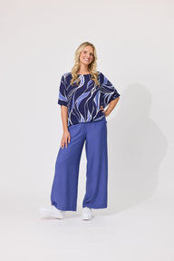 A woman modeling a short-sleeve top with a navy, white, and blue abstract wave pattern, paired with solid blue wide-leg pants and white sneakers, against a plain white studio background.