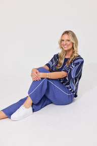 A woman sitting on a white studio floor wearing a blue and white patterned short-sleeve top and matching blue pants with white piping, paired with white sneakers.