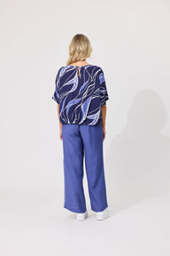 A woman viewed from the back wearing a navy blue patterned short-sleeve top with abstract white and light blue lines, paired with wide-leg blue pants and white shoes, against a plain light gray studio background.
