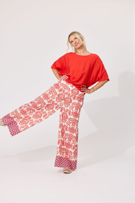 A woman posing in a bright orange short-sleeve top and wide-leg pants featuring a red and white floral pattern with solid red cuffs, set against a plain white studio background.