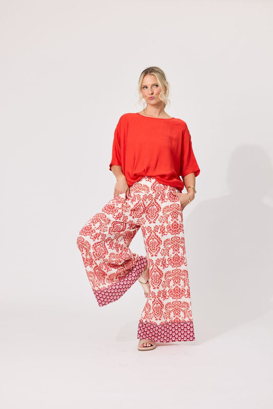 A woman modeling a bright orange short-sleeve top and wide-leg pants with a red and white floral pattern, standing against a plain light gray studio background.