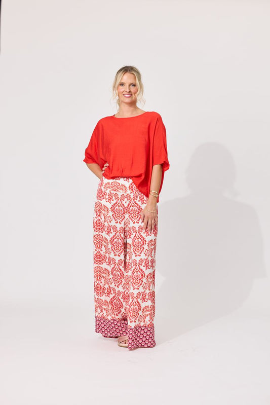 A woman modeling a bright orange short-sleeve top and wide-leg trousers with a red and white damask pattern, standing against a plain white studio background.