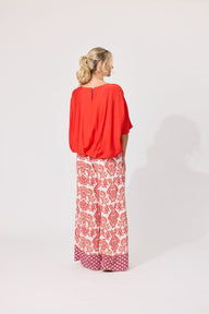A woman is shown from the back wearing a bright orange top and wide-leg pants with a red and white floral pattern, standing against a plain light gray studio background.