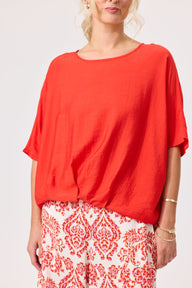 A woman wearing a loose, bright orange short-sleeve top and white pants with a red damask pattern, photographed against a light gray studio background.