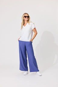 A woman modeling a white V-neck short-sleeve top and wide-leg blue pants against a plain light gray studio background.