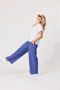 A woman wearing blue wide-leg sweatpants, a white t-shirt, and white sneakers poses in a studio setting against a light gray background.