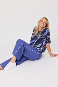 A woman wearing a blue and white abstract patterned short-sleeve top and matching blue trousers with white piping, sitting on a white studio floor.