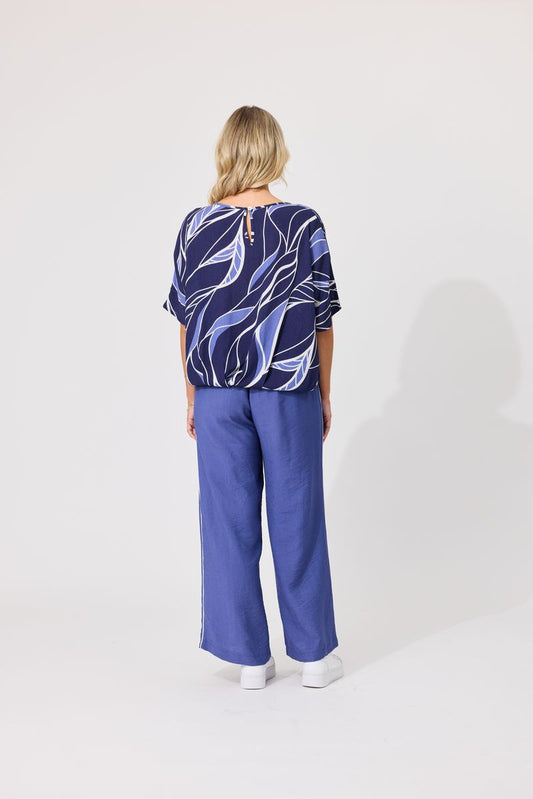 A woman viewed from the back wearing a navy blue patterned short-sleeve top with abstract white and light blue lines, paired with wide-leg blue pants and white shoes, against a plain light gray studio background.