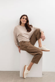 Woman sitting on a white cube wearing a beige sweater and brown pants against a white background