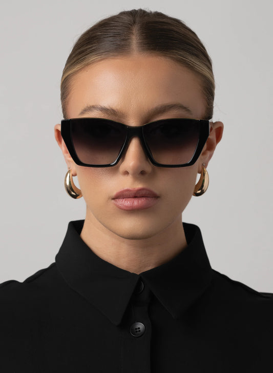 Woman wearing black sunglasses and gold hoop earrings against a gray background