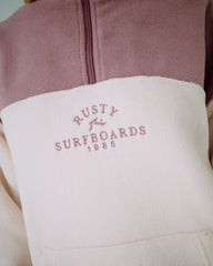 Close-up of a jacket with 'Rusty Surfboards 1985' logo on a pink and beige background