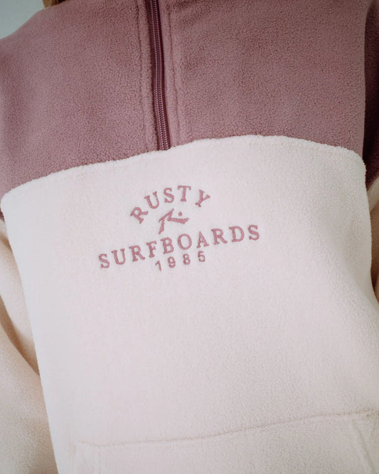 Close-up of a jacket with 'Rusty Surfboards 1985' logo on a pink and beige background