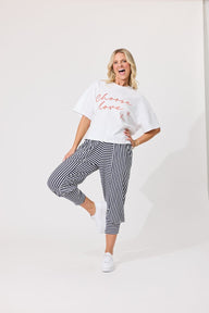 A woman modeling a white graphic t-shirt with red cursive text and black-and-white vertically striped cropped pants, posing in a bright studio setting.