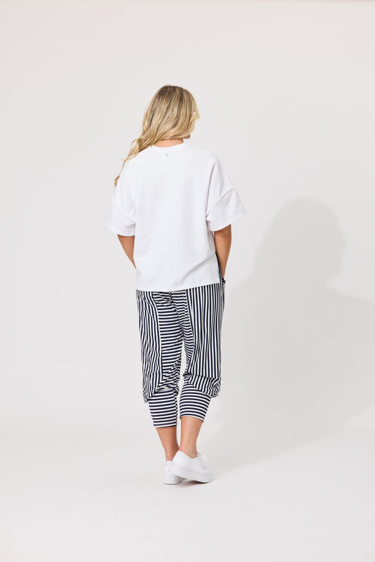 A woman viewed from behind wearing a white short-sleeve t-shirt, navy and white vertically striped cropped pants, and white sneakers, standing against a plain light gray studio background.