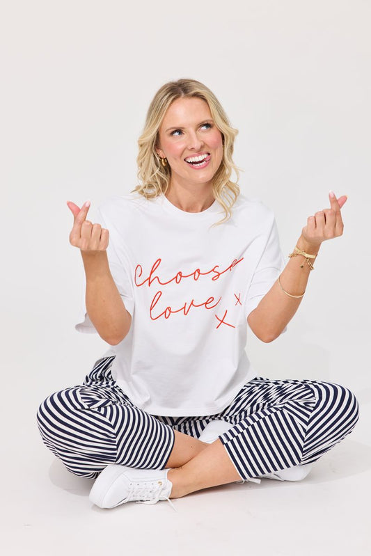 A woman sitting cross-legged on a white studio background, wearing a white graphic T-shirt with red script text reading 'choose love x' and black-and-white striped pants.