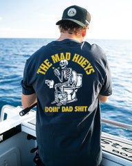 A man on a boat seen from behind wearing a black t-shirt with a large back graphic. The graphic features a white skeleton sitting on a toilet holding a beer, with yellow text reading 'THE MAD HUEYS' above and 'DOIN' DAD SHIT' below.