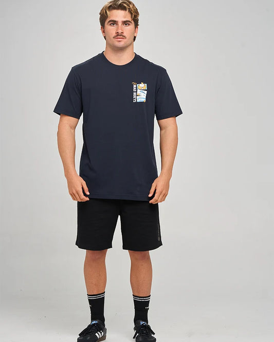 A man modeling a navy blue short-sleeve t-shirt with a small graphic on the left chest, paired with black athletic shorts, black socks, and black sneakers, against a light gray studio background.