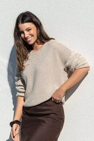 A woman posing outdoors against a white wall, wearing a beige knit sweater and a dark brown midi skirt, with white strappy heels.