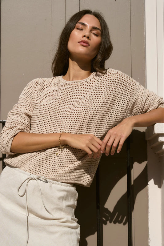 Woman wearing a beige knitted sweater and white pants against a neutral background