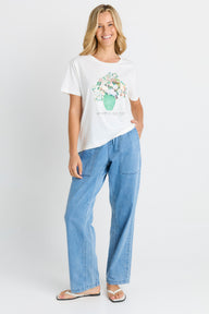 A woman modeling a white short-sleeve graphic T-shirt with a floral print and light blue wide-leg jeans against a plain light gray studio background.