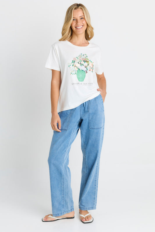 A woman modeling a white short-sleeve graphic T-shirt with a floral print and light blue wide-leg jeans against a plain light gray studio background.