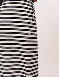 Close-up of a black and white horizontally striped garment, likely a skirt or dress, with a small white square patch featuring a black cross on the front, set against a plain off-white background.