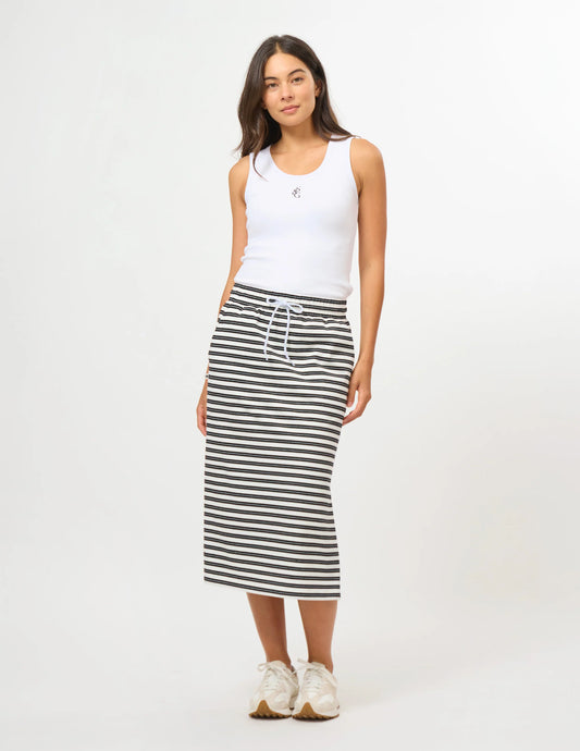 A woman modeling a white ribbed tank top and a black and white horizontally striped midi skirt with a white drawstring, paired with white sneakers, against a plain white studio background.